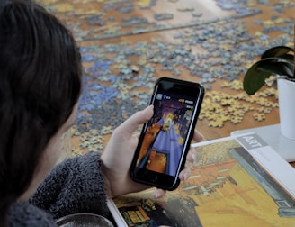 A person is playing a game on a smartphone. In the foreground, there is a puzzle spread out on the table with some pieces assembled, and a booklet featuring artwork. A plant with green leaves is placed on the table.